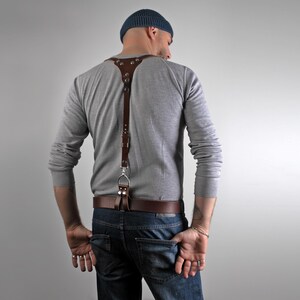May include: A brown leather suspender with silver-tone hardware. The suspender is attached to a brown leather belt. The person is wearing a gray long-sleeve shirt, blue jeans, and a blue beanie.