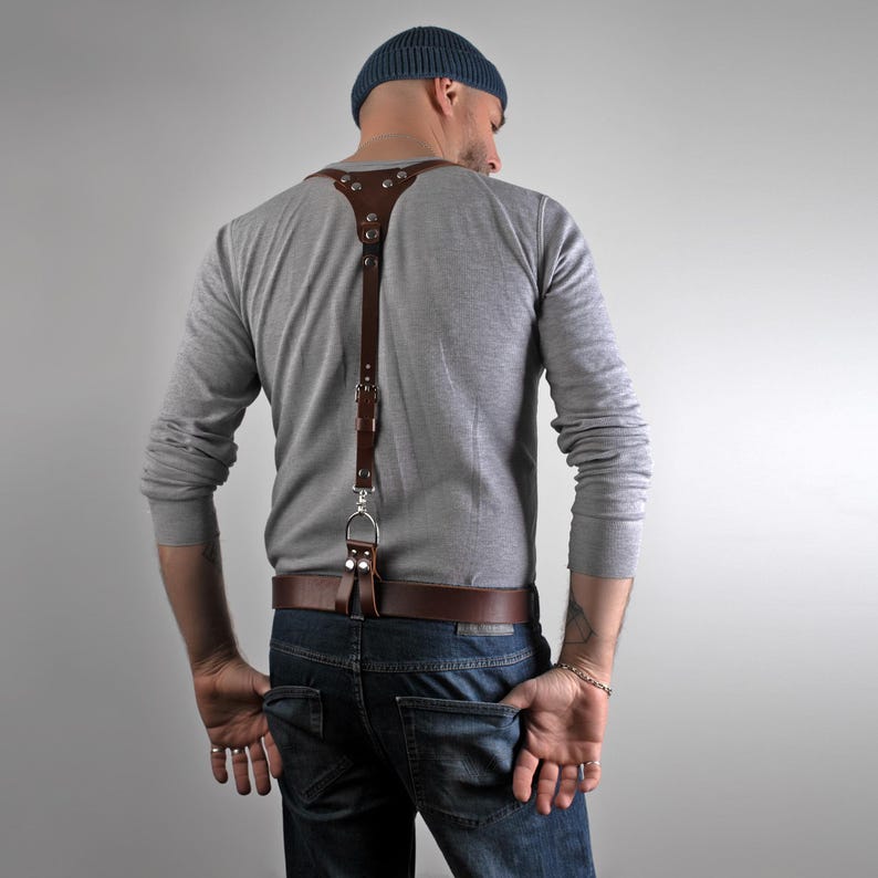 May include: A brown leather suspender with silver-tone hardware. The suspender is attached to a brown leather belt. The person is wearing a gray long-sleeve shirt, blue jeans, and a blue beanie.