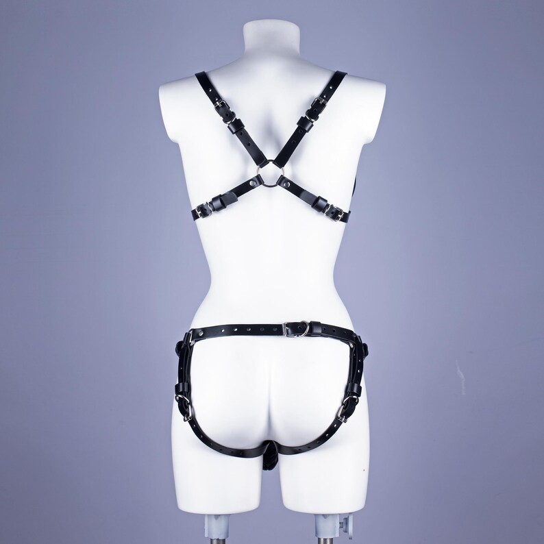 May include: Black leather body harness with adjustable straps and metal hardware. The harness has a crisscross design on the back and a separate piece around the hips. The product is displayed on a white mannequin.