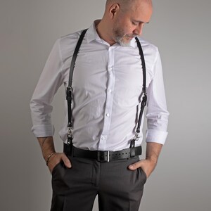 May include: Black leather suspenders with silver-tone hardware are attached to a black belt. The suspenders are worn over a white button-down shirt and gray pants. The belt has a silver buckle.