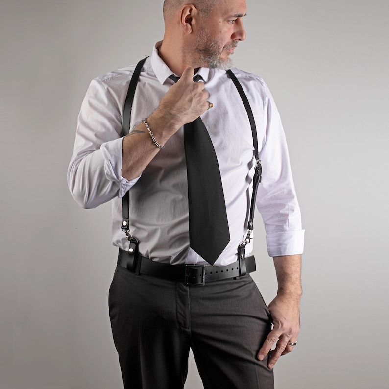 May include: A person wearing a white dress shirt, black tie, and black leather suspenders. The person is also wearing a black belt and gray dress pants. The suspenders have metal clasps.