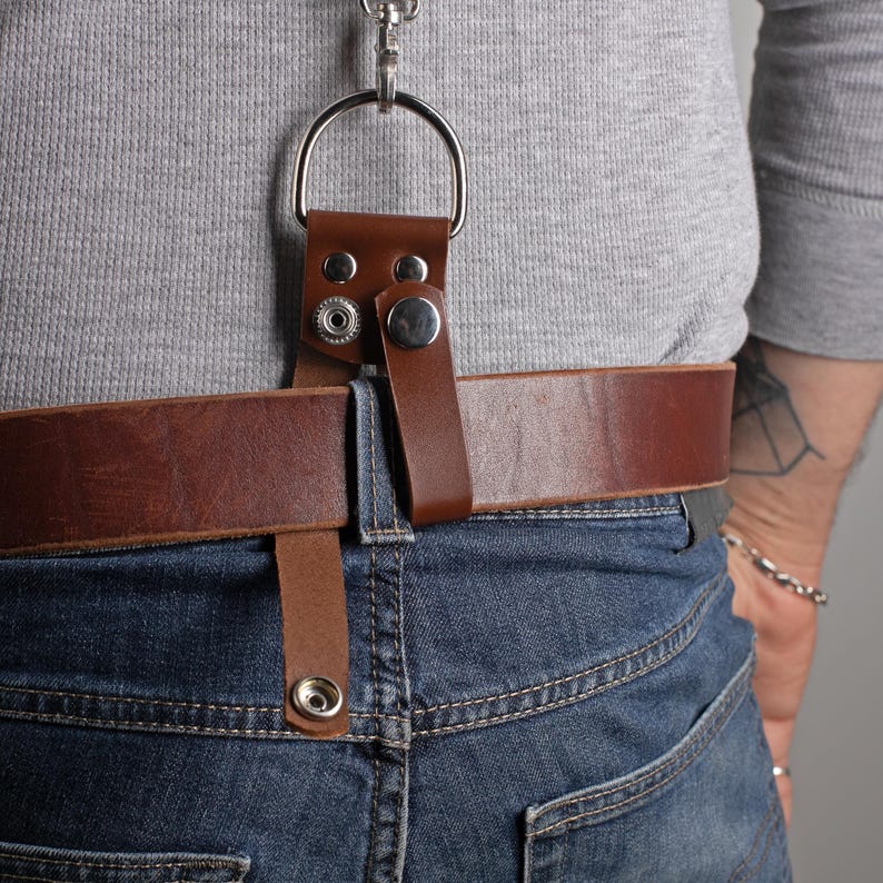 May include: A brown leather belt loop with a silver metal ring and clasp, attached to a brown leather belt. The belt is worn with blue jeans. The loop is secured with silver snap closures.