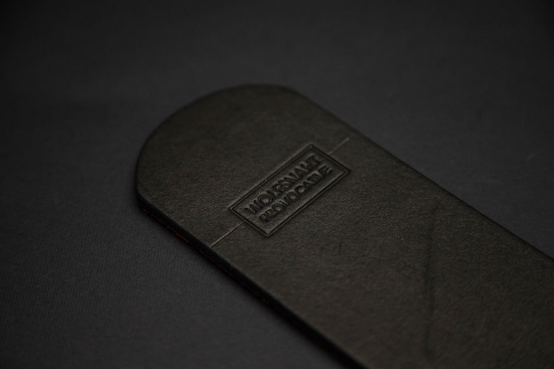 May include: A black leather item with rounded edges, featuring the embossed text "WOLFSNAKE PROVOCATIVE" in a rectangular frame. The item has a textured surface and a dark background.