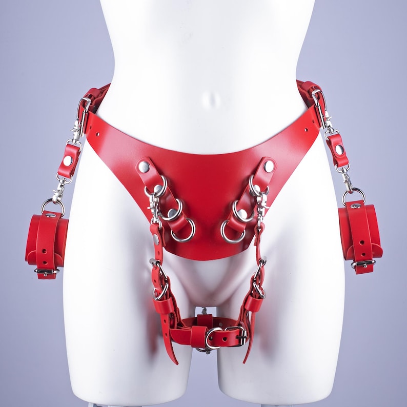 May include: A red leather harness with attached wrist restraints. The harness features multiple straps, metal rings, and silver-toned hardware. The design includes a waist belt, leg straps, and a crotch strap, all in a vibrant red color.