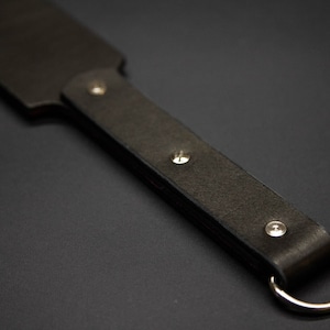 May include: A dark brown leather paddle with a rounded end and a handle. The handle is secured with three silver rivets and features a silver ring at the end. The paddle is designed for impact and is likely used for a specific purpose.