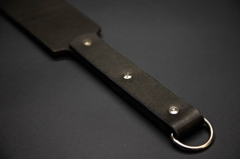 May include: A dark brown leather paddle with a rounded end and a handle. The handle is secured with three silver rivets and features a silver ring at the end. The paddle is designed for impact and is likely used for a specific purpose.