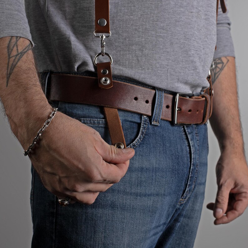 May include: Brown leather suspenders with silver hardware attached to a brown leather belt. The suspenders are clipped to the belt and jeans. The person is wearing blue jeans and a gray long-sleeve shirt.