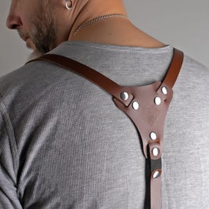 May include: Brown leather camera harness with silver rivets and a decorative emblem. The harness is designed to distribute the weight of a camera, providing comfort and stability for photographers.