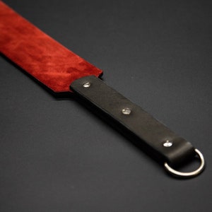 May include: A leather paddle with a dark red suede striking surface and a dark brown handle. The handle has a silver-colored ring and is secured with silver rivets. The item is set against a dark background.