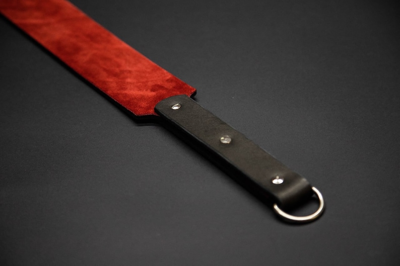 May include: A leather paddle with a dark red suede striking surface and a dark brown handle. The handle has a silver-colored ring and is secured with silver rivets. The item is set against a dark background.