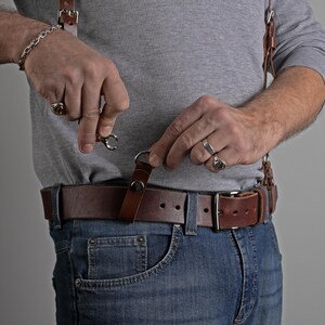 May include: Brown leather suspenders with metal clasps attached to a brown leather belt. The belt is worn with blue jeans. The suspenders are being attached to the belt.