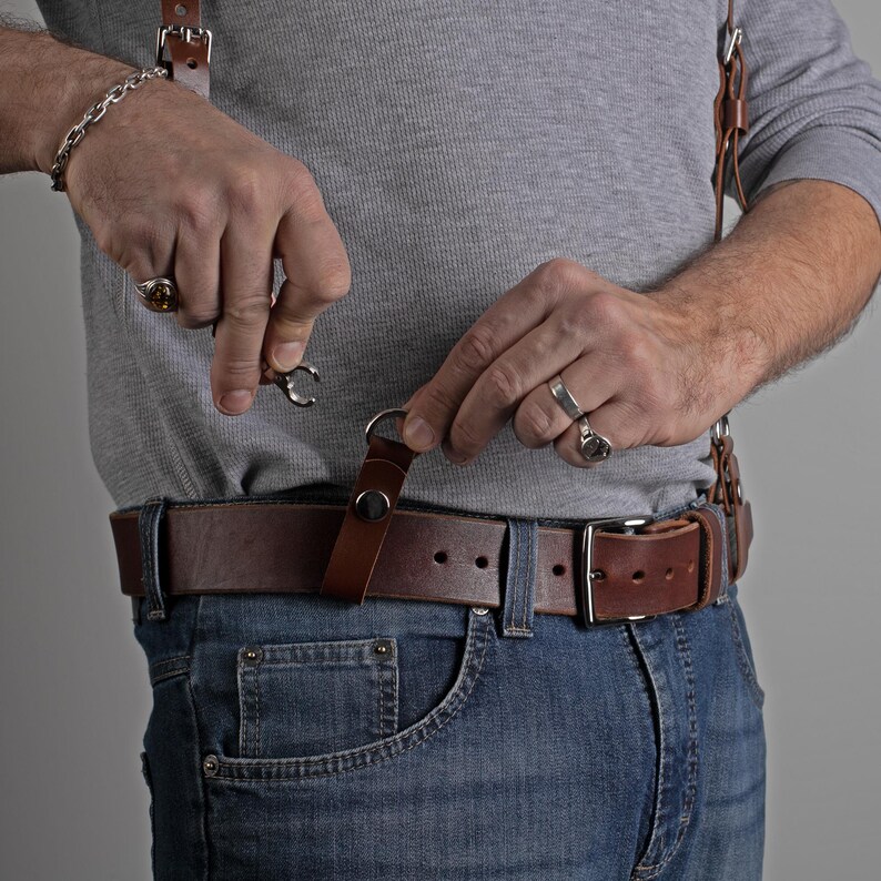 May include: Brown leather suspenders with metal clasps attached to a brown leather belt. The belt is worn with blue jeans. The suspenders are being attached to the belt.