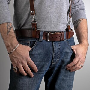 May include: Brown leather belt with a silver buckle and matching suspenders. The suspenders attach to the belt with silver clips. The person is wearing blue jeans and a gray long-sleeve shirt.