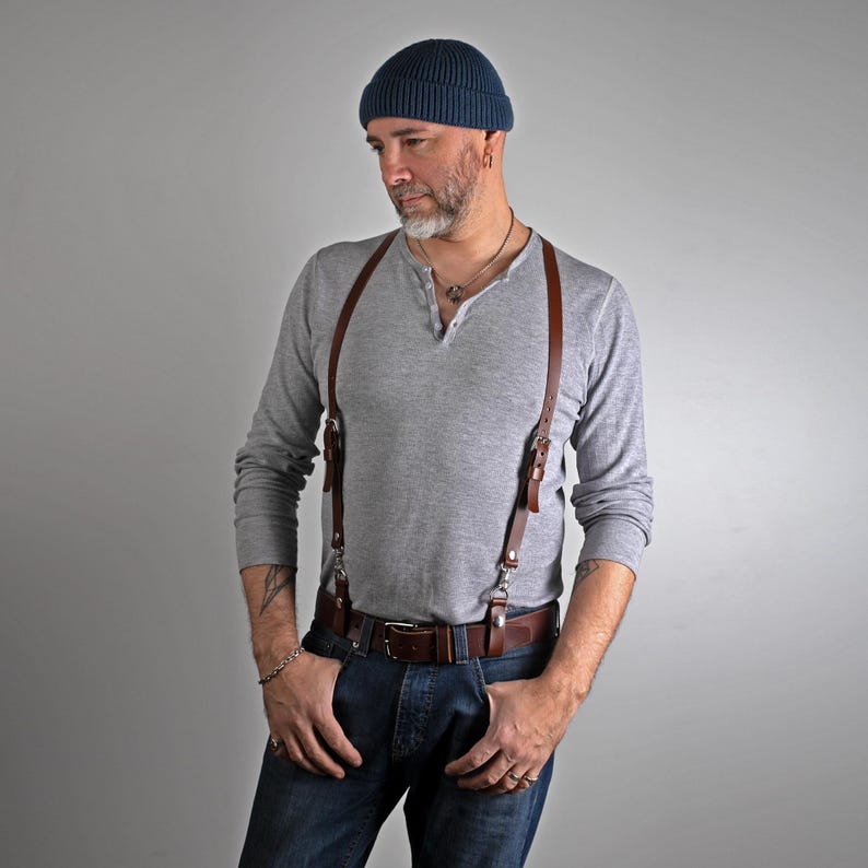 May include: Brown leather suspenders with silver-tone hardware. The suspenders are attached to a brown leather belt with a silver buckle. The person is wearing a gray long-sleeve shirt, blue jeans, and a blue beanie.
