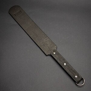 May include: A black leather paddle with a rectangular striking surface and a rounded handle. The handle is secured with silver rivets and a metal ring. The paddle has a textured surface and a small embossed logo.