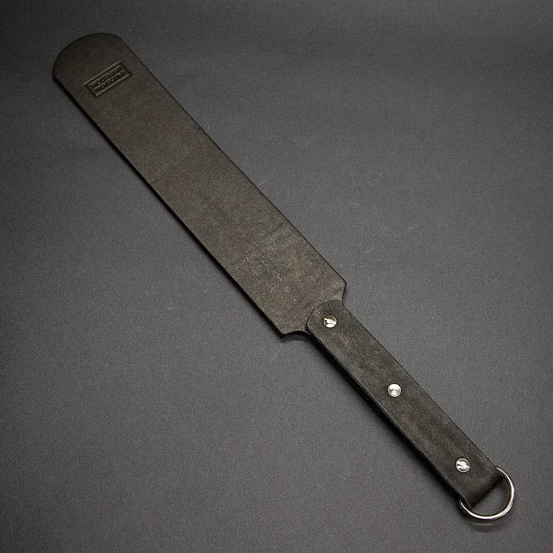 May include: A black leather paddle with a rectangular striking surface and a rounded handle. The handle is secured with silver rivets and a metal ring. The paddle has a textured surface and a small embossed logo.