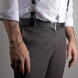 May include: A silver chain bracelet adorns a wrist, complemented by a ring. The person is wearing a white dress shirt, gray trousers, a black belt with a silver buckle, and black suspenders.