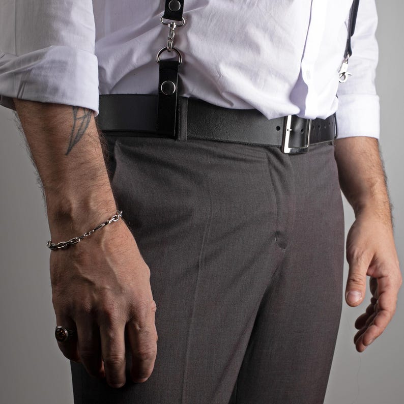 May include: A silver chain bracelet adorns a wrist, complemented by a ring. The person is wearing a white dress shirt, gray trousers, a black belt with a silver buckle, and black suspenders.
