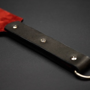 May include: A close-up of a leather paddle with a red suede striking surface. The dark brown handle is secured with silver rivets and a silver ring. The paddle is designed for impact.