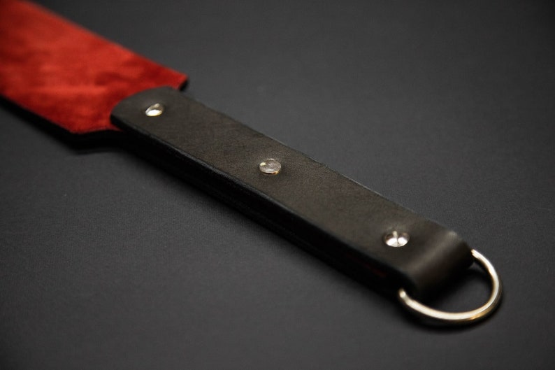 May include: A close-up of a leather paddle with a red suede striking surface. The dark brown handle is secured with silver rivets and a silver ring. The paddle is designed for impact.