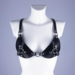 May include: Black leather harness bra with adjustable straps and silver-tone metal hardware. The bra features a unique design with circular metal rings and rivets, displayed on a white mannequin. The bra is designed to be worn as a top.