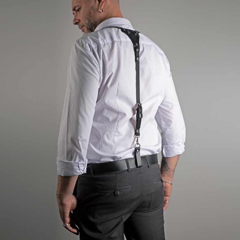 May include: Black leather suspenders with silver-tone hardware. The suspenders attach to the shoulders and waist with adjustable straps and a metal clasp. The suspenders are worn over a white dress shirt and gray trousers.