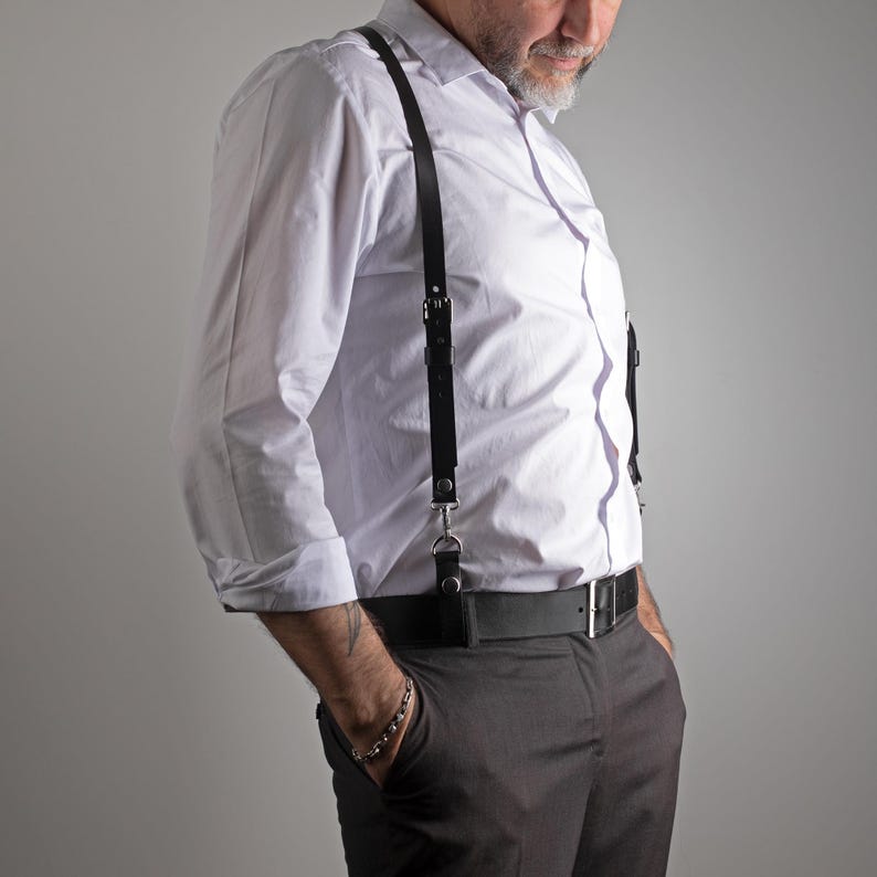May include: Black leather suspenders with silver-tone hardware. The suspenders are attached to a black belt and paired with gray trousers and a white button-down shirt. The suspenders have adjustable straps and a classic design.