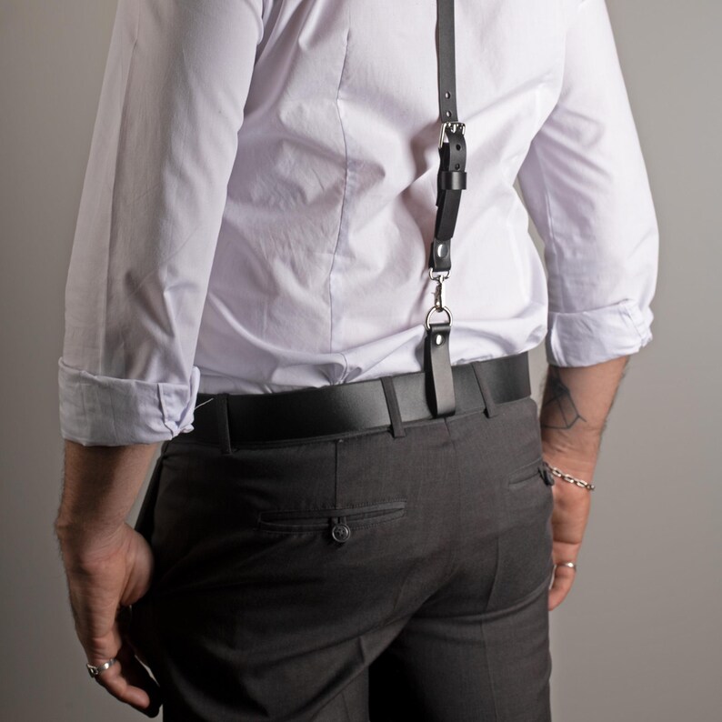 May include: Black leather suspender with a silver-tone clasp attached to a black belt. The suspender is worn over a white dress shirt and gray trousers. The shirt sleeves are rolled up.