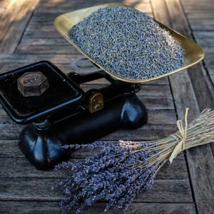 Könnte beinhalten: Eine schwarze mechanische Vintage-Waage mit einer Messingschale, die mit getrockneten violetten Lavendelblüten gefüllt ist. Ein dunkles Metallgewicht liegt auf der gegenüberliegenden Plattform. Neben der Waage liegt ein mit Bindfaden gebundenes Lavendelbündel.