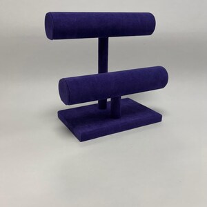 May include: A purple velvet jewellery display stand with two horizontal bars and a rectangular base. The bars are cylindrical and supported by vertical posts. The stand is designed to showcase bracelets or necklaces. The background is plain white.