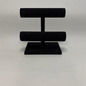 May include: Black velvet jewellery display stand with two horizontal bars. The stand has a square base and two vertical supports. The bars are designed to hold bracelets, watches, or necklaces. The stand is designed to showcase jewellery.