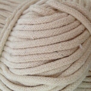 May include: A close-up of a large spool of natural-colored cotton rope. The rope is tightly wound and has a braided texture.