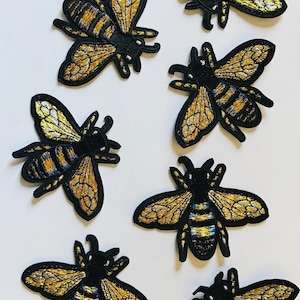 May include: Seven gold and black embroidered bee patches. The bees have black bodies, gold wings, and blue and gold stripes.