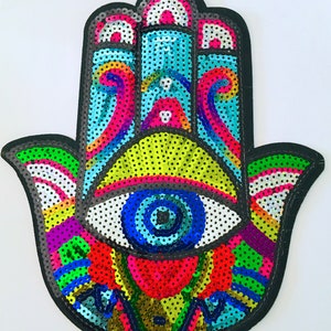 May include: A colorful sequined Hamsa hand patch with a black border. The design features an eye in the center, surrounded by vibrant hues of blue, green, yellow, red, and purple. The patch is likely for decorative purposes.