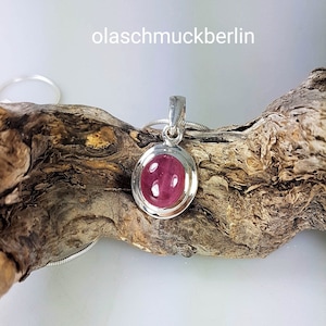 May include: A silver pendant with a round pink gemstone set in a bezel setting. The pendant is on a silver chain and is resting on a piece of driftwood.