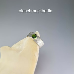 May include: A silver ring with a textured finish and a green gemstone set in a gold bezel. The ring is displayed on a white rock.