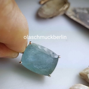 May include: A light blue aquamarine gemstone set in a silver pendant. The pendant is held in a hand, and the background is a white surface.