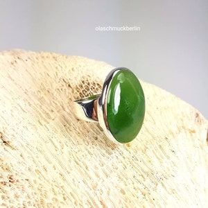 May include: A silver ring with a green oval gemstone. The ring is set on a light brown surface.