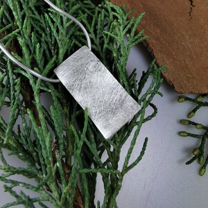 Puede incluir: Un collar de plata con un colgante rectangular texturizado. El colgante tiene un acabado cepillado y cuelga de una cadena de plata.