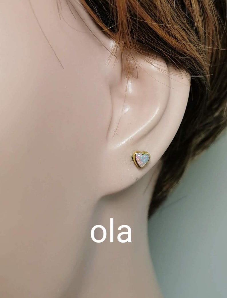 May include: A pair of gold heart-shaped earrings with opal stones. The earrings are shown on a model's ear.