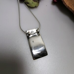 Puede incluir: Un collar de plata con un colgante rectangular. El colgante es liso y brillante, y tiene una forma ligeramente curvada.