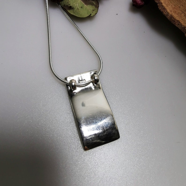 Puede incluir: Un collar de plata con un colgante rectangular. El colgante es liso y brillante, y tiene una forma ligeramente curvada.
