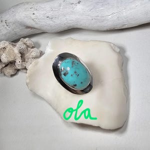 May include: A silver ring with an oval turquoise stone. The turquoise stone is a vibrant blue-green color with dark spots. The ring is set on a white surface with a piece of driftwood in the background.