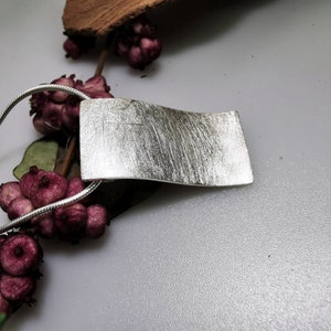 Puede incluir: Un collar de plata con un colgante de acabado cepillado. El colgante es de forma rectangular con bordes redondeados y una ligera curva. El collar est&aacute; en una cadena de plata.