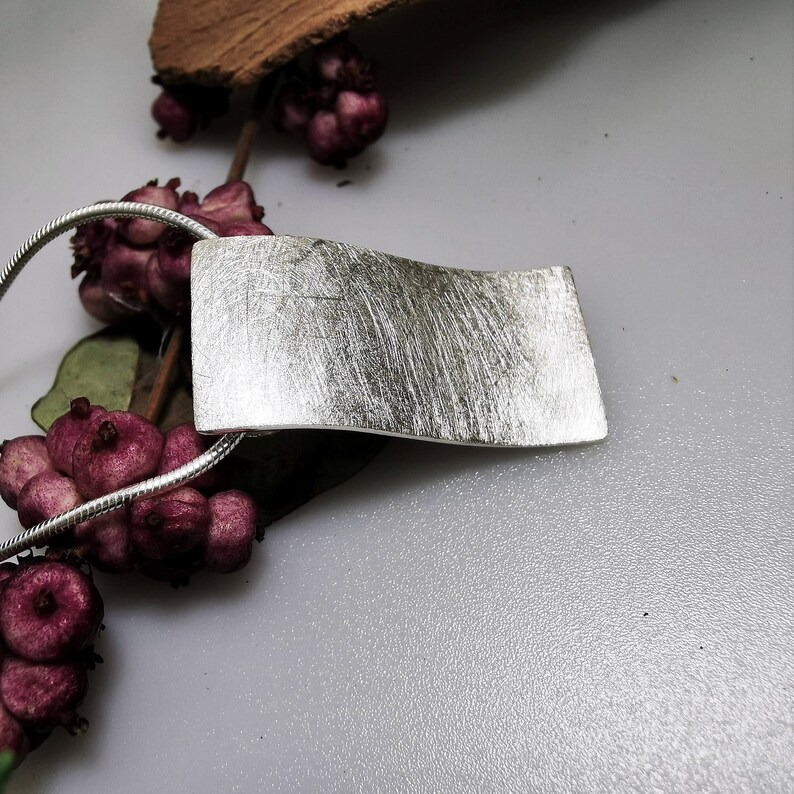 Puede incluir: Un collar de plata con un colgante de acabado cepillado. El colgante es de forma rectangular con bordes redondeados y una ligera curva. El collar est&aacute; en una cadena de plata.