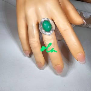 May include: A silver ring with a large, oval, bright green gemstone. The ring has a textured, organic design around the stone. The ring is displayed on a hand with manicured nails.