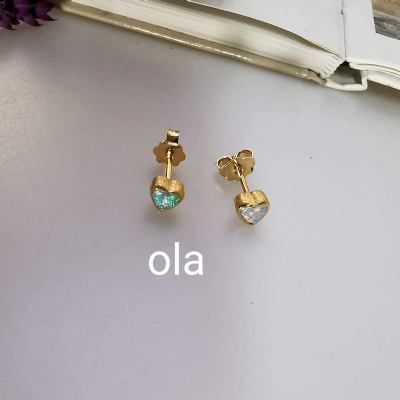 May include: A pair of gold heart-shaped earrings with opal stones. The earrings are on a white surface with the word "ola" written below them.