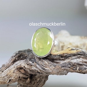 May include: A silver ring with a large, oval-shaped, light green gemstone. The ring is set on a wooden surface.