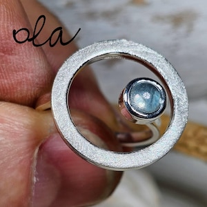 Puede incluir: Un anillo de plata con un diseño circular texturizado. El anillo presenta una pequeña gema redonda de color azul claro engastada en el círculo. La palabra "ola" está escrita en cursiva.