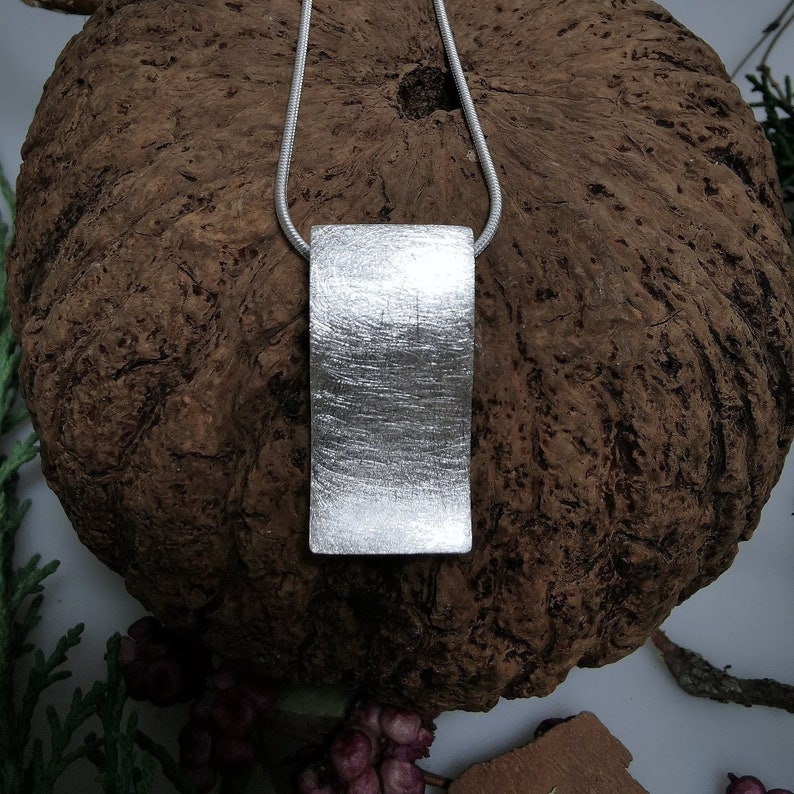 Puede incluir: Un collar con un colgante de plata cepillado. El colgante es rectangular y ligeramente curvado. El collar est&aacute; en una cadena de plata.